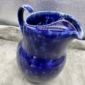 May include: A blue ceramic pitcher with a white marbled glaze. The pitcher has a wide, rounded base and a spout with a curved lip.