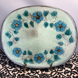 May include: A light blue ceramic serving plate with a raised rim and a decorative design of blue flowers and green leaves around the edge. The plate has a speckled glaze and is displayed on a black and white patterned surface.