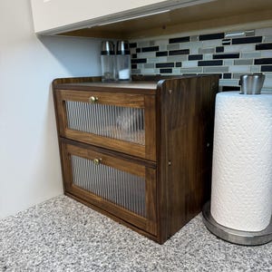 May include: A two-drawer wooden cabinet with ribbed glass doors and brass knobs. The cabinet is stained a dark brown and sits on a granite countertop. Salt and pepper shakers are on top of the cabinet.