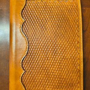 May include: A handcrafted, tan leather journal with a decorative diamond pattern and scalloped edge. The cover is stitched with white thread, and the leather has a rich, warm tone. The journal is rectangular and appears to be closed.