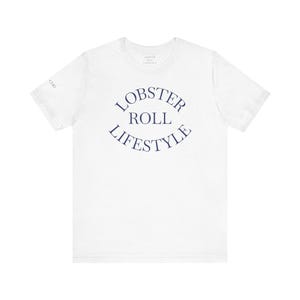 May include: White t-shirt with the text "LOBSTER ROLL LIFESTYLE" in navy blue, arched across the front. The shirt has short sleeves and a crew neck. The words "LOBSTER ROLL LIFESTYLE" are in a curved font.