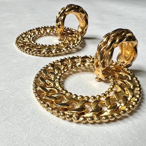 Könnte beinhalten: Ein Paar goldfarbene Creolen. Jede Ohrring hat ein großes, kreisförmiges Design mit einem kettenartigen Muster am äußeren Rand und einem geflochtenen Oberteil. Die Ohrringe werden auf hellem Hintergrund präsentiert.