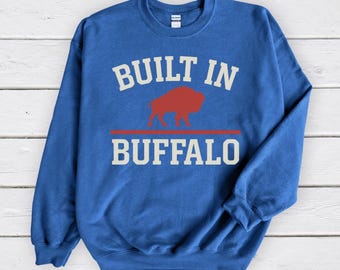 May include: A blue sweatshirt with the text "BUILT IN BUFFALO" in white block letters. A red buffalo graphic is centered between the words, above a red horizontal line. The sweatshirt is a solid blue color.