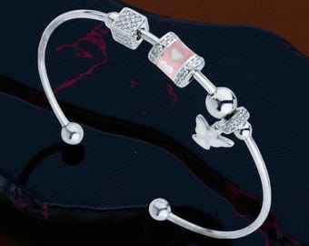 May include: Silver-toned bangle bracelet with decorative charms. The bracelet features a pink and white enamel charm with a heart design, a butterfly charm, and square charms with clear stones. The bracelet has a curved design with a ball at each end.