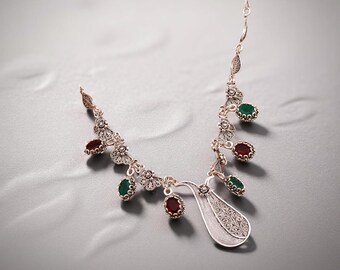 May include: A silver necklace with a delicate filigree design. The necklace features alternating red and green gemstone accents, and a teardrop-shaped pendant. The necklace is displayed on a gray surface.