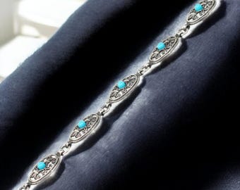 Puede incluir: Un brazalete de plata con eslabones ovalados adornados con diseños de filigrana y una pequeña piedra turquesa redonda en el centro de cada eslabón. El brazalete se muestra sobre una superficie azul oscuro.