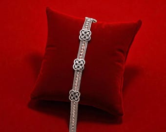 May include: A silver bracelet with three black and white flower-shaped accents. The bracelet is displayed on a red velvet pillow against a red background. The bracelet has a chain-like design.