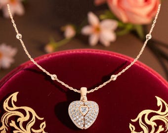 Puede incluir: Un collar dorado con un colgante en forma de corazón. El colgante está incrustado con pequeñas piedras preciosas transparentes y presenta una gema más pequeña en forma de corazón en el centro. La cadena del collar tiene pequeñas cuentas redondas.
