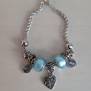 May include: Silver-toned bracelet featuring a braided chain design. The bracelet includes light blue glass beads, decorative metal charms, and a heart-shaped pendant with a cross. The bracelet is designed for a delicate and feminine aesthetic.