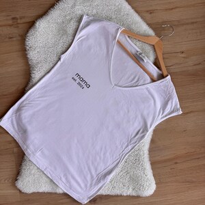 May include: A white vest top with a V-neck. The text "mama est. 2024" is printed in black on the front. The top is on a wooden hanger, resting on a fluffy, white rug, set against a wooden background.