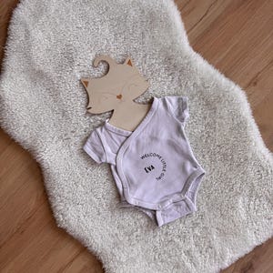 May include: A light-colored baby onesie with the text "WELCOME LITTLE GIRL EVA" is laid on a fluffy white rug. A wooden fox-shaped hanger is placed above the onesie. The background is a light-colored wooden floor.