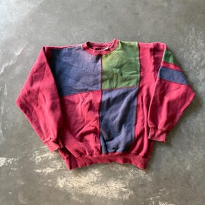 May include: A long-sleeve crewneck sweatshirt featuring a color-blocked design. The garment is primarily red with large rectangular panels in muted blue and olive green on the front chest area, and additional blue and green stripes on the sleeve.