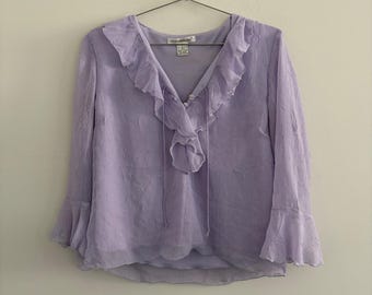 May include: A light purple long-sleeve blouse featuring a V-neckline with a ruffled collar, two small buttons at the center, and thin tie strings. The fabric has a subtle crinkled texture, and the sleeves end in flared, ruffled cuffs.