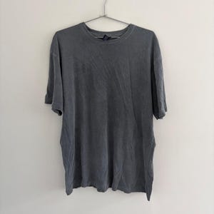 Pode incluir: Uma t-shirt de manga curta com gola redonda num tom cinzento-carvão sólido com um aspeto lavado e desbotado. A peça apresenta uma bainha reta padrão, uma gola canelada e está pendurada num cabide de metal contra uma parede lisa.
