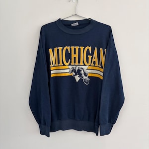 Può includere: Una felpa girocollo blu navy con la scritta MICHIGAN in grandi lettere gialle. Sotto il testo, un triangolo bianco contiene un'illustrazione grafica di un ghiottone, affiancato da due strisce orizzontali gialle sul petto.