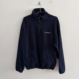 May include: A navy blue fleece pullover featuring a quarter-zip closure with a high collar. The garment includes long sleeves, elastic cuffs, and a front kangaroo pocket. The word CHAPS is embroidered in white on the left chest area.