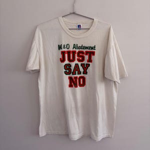 May include: A white short-sleeve t-shirt featuring the text M&O Abatement and JUST SAY NO in red block letters with green outlines. The shirt has a crew neckline and a Russell brand tag visible at the inner collar, hanging on a metal hanger.