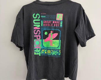 May include: A charcoal gray short-sleeve t-shirt featuring a vibrant graphic on the back. The design includes the text SUNSPORT, GREAT WAVE BUGLE BOY, and THE BEGINNING OF THE ADVENTURE in neon green and pink, with a stylized surfer silhouette.