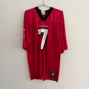 May include: A red short-sleeved jersey with a black V-neck collar. The front features the text FALCONS above a large white number 7 with a black outline. A small NFL logo patch is at the bottom left, and a Reebok logo is on the right sleeve.