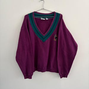 Könnte beinhalten: Ein dunkelviolettes Langarm-Sweatshirt mit V-Ausschnitt und gestreifter Verzierung in Petrol und Grün. Das Kleidungsstück verfügt über ein kleines gesticktes Wappenlogo auf der linken Brustseite und hängt an einem Metallbügel vor einer Wand.