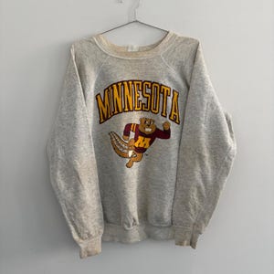 Könnte beinhalten: Ein hellgrau meliertes Sweatshirt mit Rundhalsausschnitt und dem Wort MINNESOTA in großen gelben Buchstaben mit dunkelroter Umrandung. Unter dem Text befindet sich eine Grafik eines Zeichentrick-Gophers in einem weinrot-goldenen Trikot mit gelbem M.