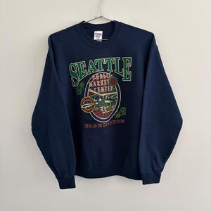 May include: A navy blue crewneck sweatshirt featuring a graphic print that reads SEATTLE PUBLIC MARKET CENTER WASHINGTON. The design includes green, red, and white illustrations of the market area, including a fish and a clock, on the chest.
