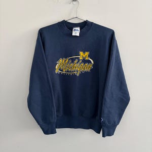May include: A navy blue crewneck sweatshirt featuring the yellow and white embroidered text Michigan Wolverines with a large yellow block M logo above it. The garment has long sleeves, ribbed cuffs, and a Pro Player brand tag at the neckline.