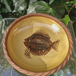 Könnte beinhalten: Ein Keramikteller mit Fischmuster. Der Teller hat eine gelbe Mitte mit einer braunen Fischillustration. Der Rand ist braun mit einer seilartigen Textur. Der Teller befindet sich vor einem Hintergrund aus grünen Blättern.