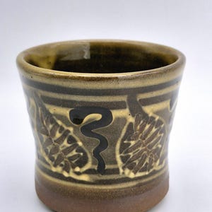May include: A handcrafted ceramic cup with a brown base and a light brown top. The cup features a repeating pattern of leaves and a black, stylized snake design. The cup is approximately 10 cm tall.
