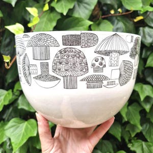 Puede incluir: Un cuenco de cerámica blanco con un diseño de setas negras. El cuenco, de forma redondeada, está sostenido por una mano. Los diseños de setas varían en estilo, con patrones detallados. El fondo es un exuberante follaje verde.