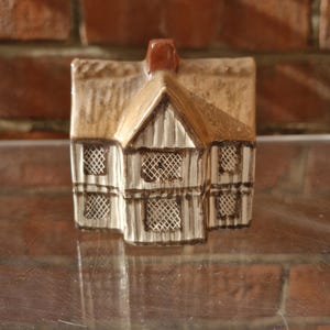 Puede incluir: Una casa de cerámica en miniatura con tejado de paja y chimenea. La casa presenta una fachada blanca y marrón con patrones de ventanas detallados. El tejado es de color marrón claro y la chimenea es marrón oscuro. Un artículo decorativo.