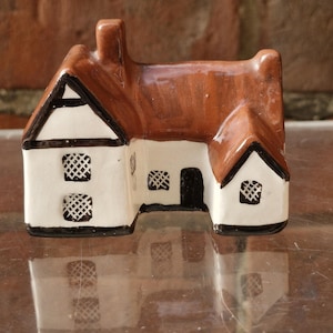 Puede incluir: Una casa en miniatura de cerámica con tejado y chimenea marrones, paredes blancas y ribetes negros. La casa tiene dos ventanas con diseño de celosía y una puerta negra. Es un objeto decorativo.