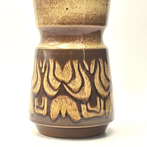 Könnte beinhalten: Eine braune Keramikvase mit strukturierter Oberfläche und einzigartigem Design. Der obere Teil ist hellbraun, während der mittlere Teil ein sich wiederholendes Muster in einer kontrastierenden Goldfarbe aufweist. Die Vase hat eine breite, zylindrische Form.