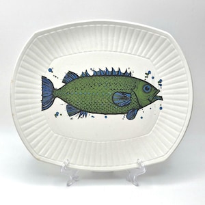 May include: White ceramic serving platter with a green and blue fish illustration. The fish has a textured body and detailed fins. The platter has a ridged border and is displayed on a clear stand.