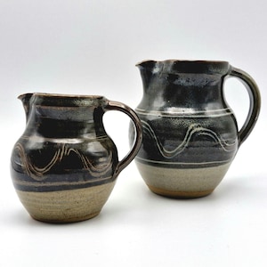 May include: Two ceramic jugs with dark brown and tan glaze. The jugs have a wavy design and a handle. The smaller jug is on the left, and the larger jug is on the right. The jugs are handmade and have a rustic appearance.