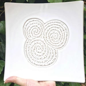 Puede incluir: Un plato cuadrado de cerámica blanca con un diseño en relieve texturizado de tres espirales interconectadas. El plato se sostiene en una mano, con follaje verde en el fondo. El diseño del plato es de color beige claro.