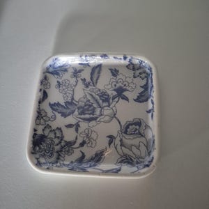 May include: Square ceramic dish with a white base and a blue floral pattern. The design features detailed flowers and leaves. The dish has rounded corners and a slightly raised edge. The overall aesthetic is classic and decorative.