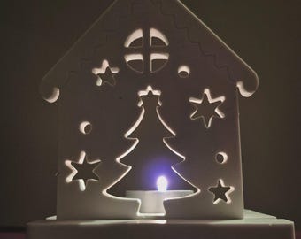 May include: A white, house-shaped candle holder with a Christmas tree and star cutouts. A small candle is lit inside, creating a warm glow. The design includes a window and decorative roof trim.