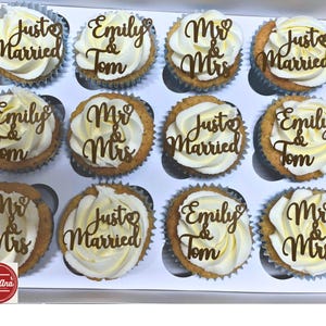 Op de afbeelding: Een doos met twaalf cupcakes met witte frosting en gouden toppers. De toppers zeggen "Just Married", "Emily & Tom" en "Mr & Mrs". De cupcakes zitten in papieren vormpjes, ideaal voor een bruiloft.