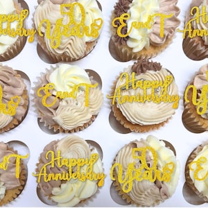 Peut inclure: Une douzaine de cupcakes avec un glaçage blanc et marron, chacun surmonté d'un décor doré scintillant "Joyeux Anniversaire" ou "50 ans". Les cupcakes sont disposés dans une boîte blanche.