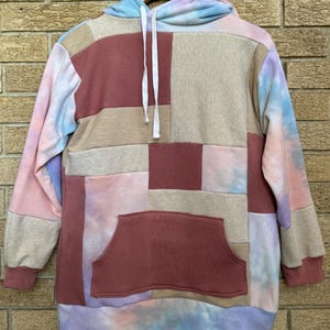 Puede incluir: Una sudadera con capucha de patchwork en tonos rosa, beige y burdeos, con efecto tie-dye en la capucha y la parte inferior. La sudadera tiene un bolsillo de canguro y un cord&oacute;n. El dise&ntilde;o es una mezcla de formas geom&eacute;tricas.