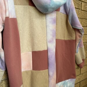 Puede incluir: Una sudadera con capucha de patchwork en colores pastel. El dise&ntilde;o presenta cuadrados rosas, beiges y malvas, con detalles tie-dye en azul, morado y rosa. La sudadera tiene una capucha con cord&oacute;n y pu&ntilde;os de canal&eacute;.