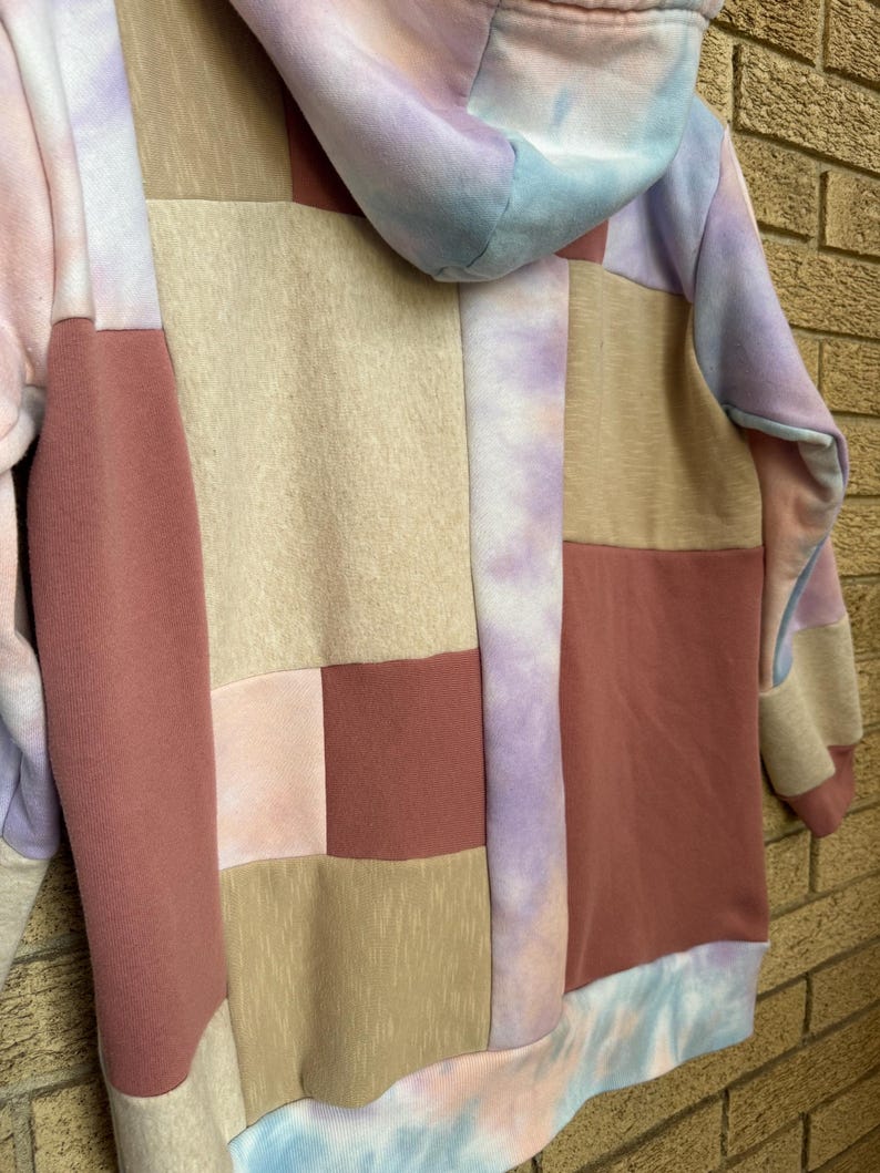 Puede incluir: Una sudadera con capucha de patchwork en colores pastel. El dise&ntilde;o presenta cuadrados rosas, beiges y malvas, con detalles tie-dye en azul, morado y rosa. La sudadera tiene una capucha con cord&oacute;n y pu&ntilde;os de canal&eacute;.