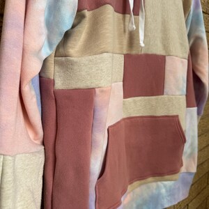 Puede incluir: Una sudadera con capucha de patchwork en tonos rosa, beige y te&ntilde;ido anudado. La sudadera tiene un bolsillo de canguro y una capucha con cord&oacute;n. Las mangas son una mezcla de te&ntilde;ido anudado y colores lisos. El dise&ntilde;o es una mezcla de texturas y colores.