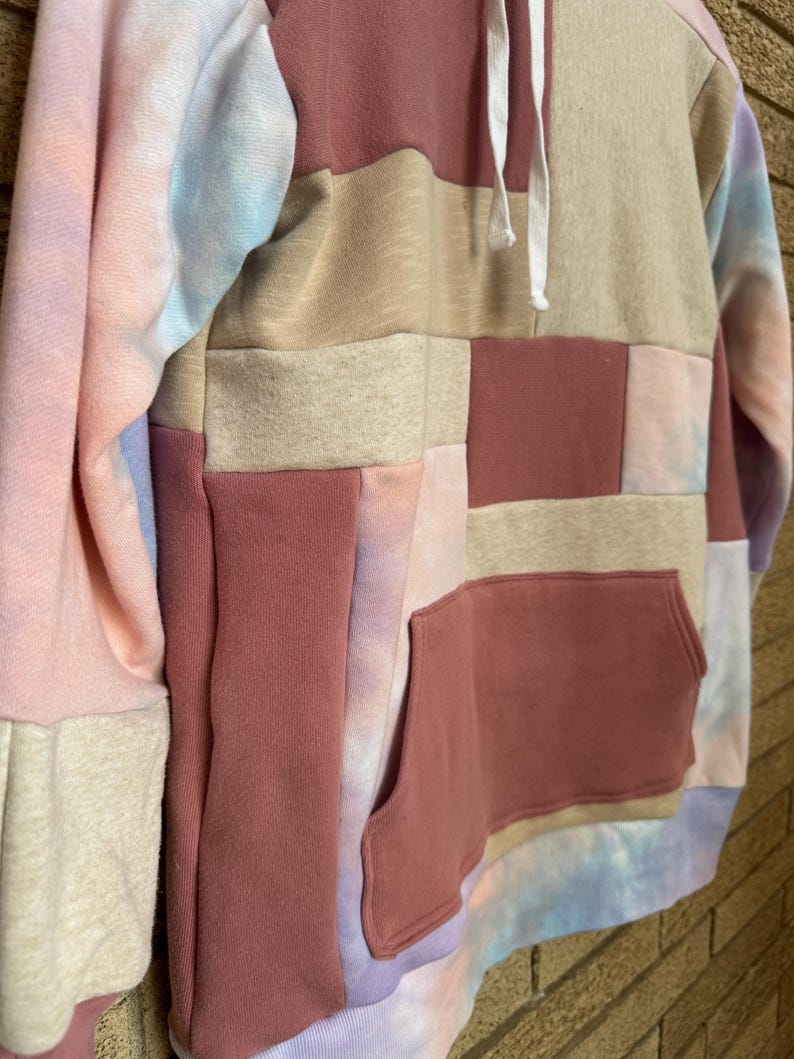 Puede incluir: Una sudadera con capucha de patchwork en tonos rosa, beige y te&ntilde;ido anudado. La sudadera tiene un bolsillo de canguro y una capucha con cord&oacute;n. Las mangas son una mezcla de te&ntilde;ido anudado y colores lisos. El dise&ntilde;o es una mezcla de texturas y colores.