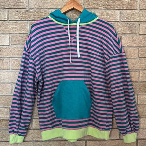 Könnte beinhalten: Ein gestreifter Hoodie in Rosa und Türkis mit einer türkisfarbenen Kapuze und Tasche. Der Hoodie hat einen Kordelzug und eine limettengrüne Einfassung an Kapuze und Bündchen. Er hängt an einem Holzkleiderbügel.