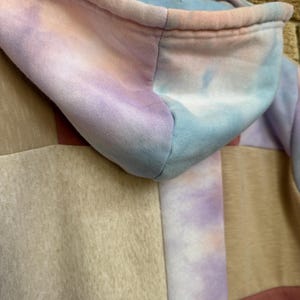 Puede incluir: Primer plano de una sudadera con capucha te&ntilde;ida con tinte anudado en colores pastel. La capucha es una mezcla de tonos azules, rosas y morados. El cuerpo de la sudadera presenta un dise&ntilde;o de retazos con paneles beige, rosas y morados. La sudadera est&aacute; colgada en una percha de madera.