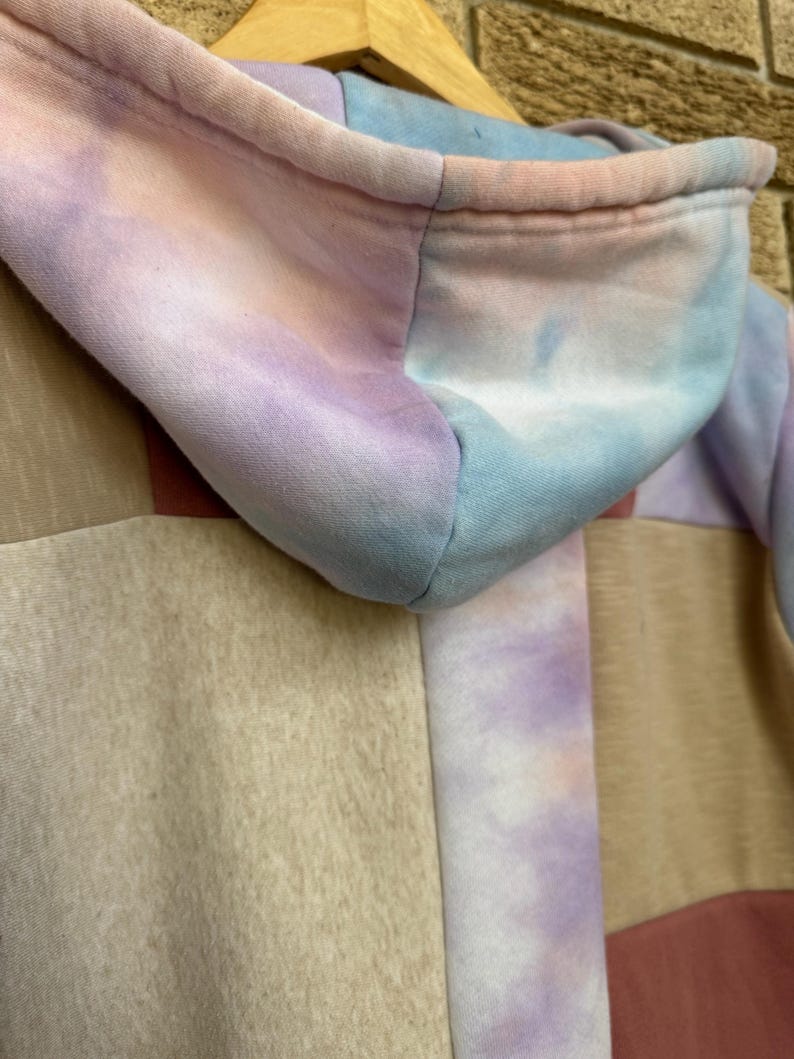 Puede incluir: Primer plano de una sudadera con capucha te&ntilde;ida con tinte anudado en colores pastel. La capucha es una mezcla de tonos azules, rosas y morados. El cuerpo de la sudadera presenta un dise&ntilde;o de retazos con paneles beige, rosas y morados. La sudadera est&aacute; colgada en una percha de madera.