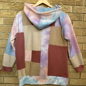 Puede incluir: Una sudadera con capucha de patchwork en tonos rosa, azul, beige y burdeos. La parte trasera de la sudadera con capucha es visible, mostrando una mezcla de paneles de tela rectangulares. La capucha y los pu&ntilde;os presentan un estampado tie-dye. La prenda est&aacute; colgada de una percha de madera.