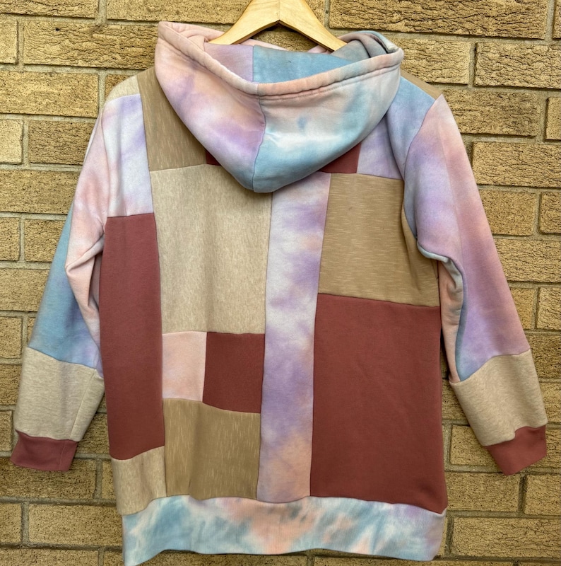Puede incluir: Una sudadera con capucha de patchwork en tonos rosa, azul, beige y burdeos. La parte trasera de la sudadera con capucha es visible, mostrando una mezcla de paneles de tela rectangulares. La capucha y los pu&ntilde;os presentan un estampado tie-dye. La prenda est&aacute; colgada de una percha de madera.
